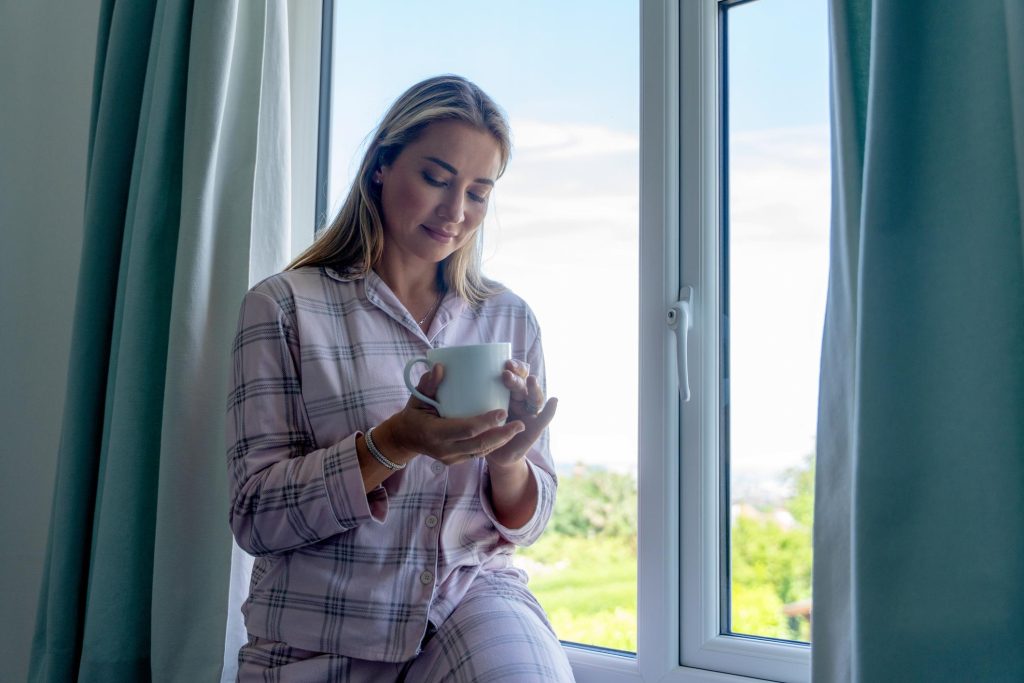 Frau genießt einen ruhigen Moment am offenen Fenster mit einer Tasse in der Hand.