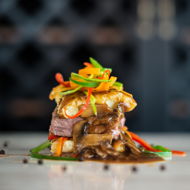 Ein gestapeltes Gericht aus Fleisch, Gemüse und einer dunklen Sauce, kunstvoll garniert.