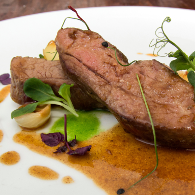 Zarte Fleischstücke auf einem Teller mit grüner Sauce und essbaren Blüten.